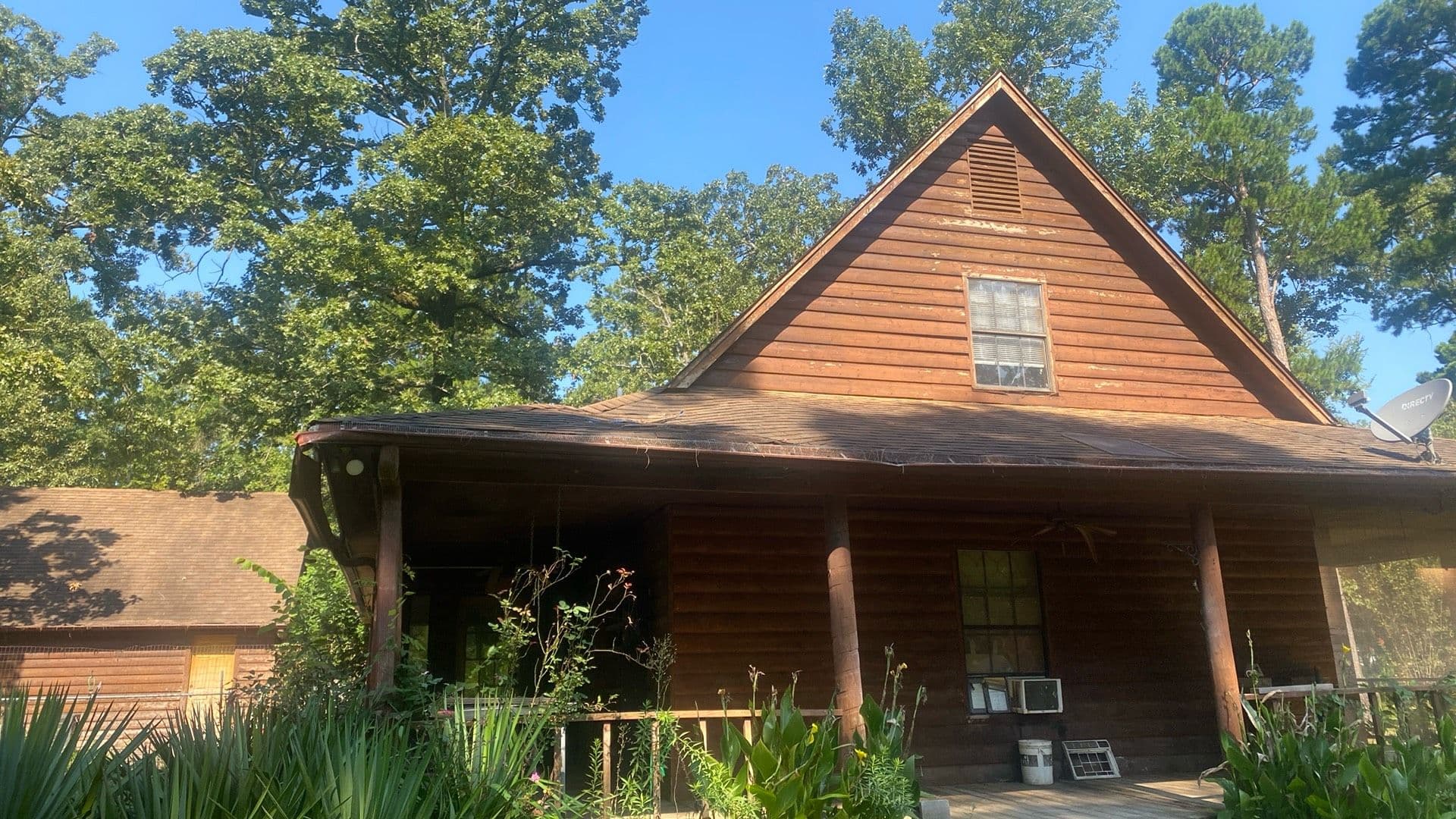 Brown wooden house surrounded by trees on a sunny day with a porch and window air conditioner.